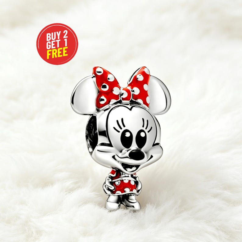 May include: A silver charm featuring Minnie Mouse with a red bow and dress, both with white polka dots. The charm has large ears and a smiling face. A red circle with the text "BUY 2 GET 1 FREE" is in the background.
