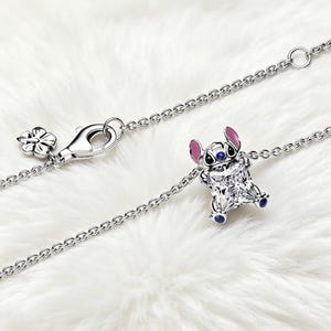 May include: A silver necklace featuring a charm of the character Stitch from the Disney movie Lilo & Stitch. The charm is a square-cut gemstone with Stitch's face and ears in blue and pink. The necklace has a flower-shaped clasp.