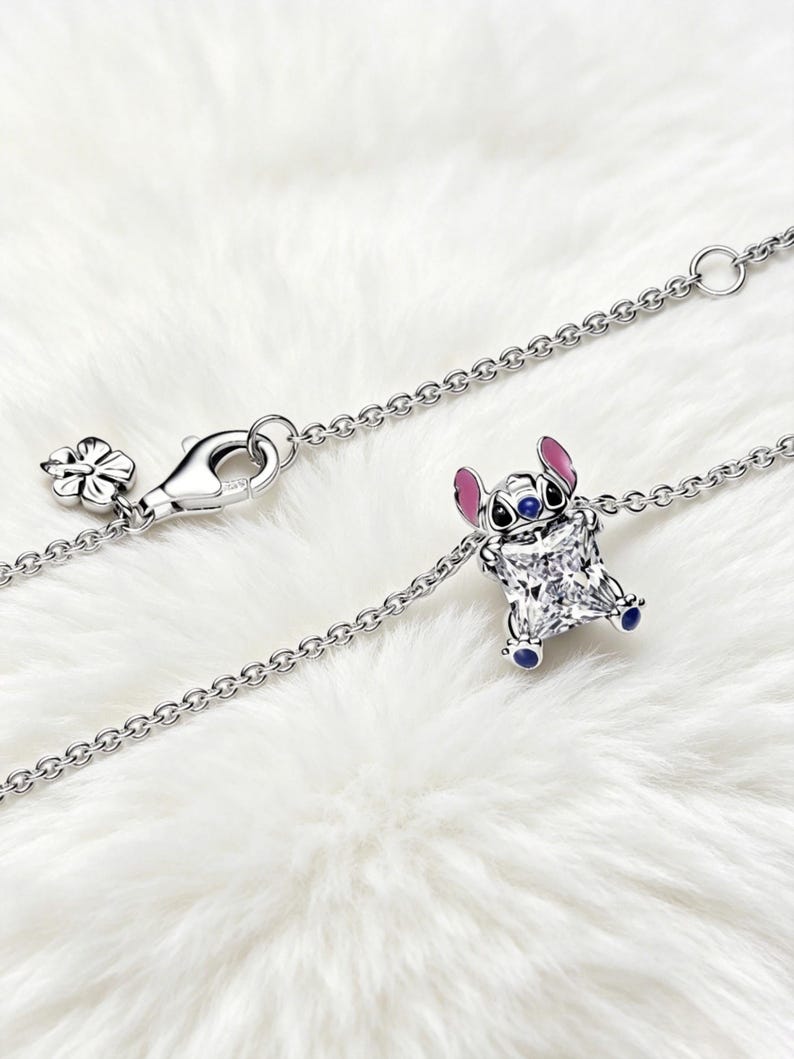 May include: A silver necklace featuring a charm of the character Stitch from the Disney movie Lilo & Stitch. The charm is a square-cut gemstone with Stitch's face and ears in blue and pink. The necklace has a flower-shaped clasp.