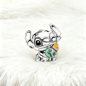 May include: A silver-colored figurine of the character Stitch from Lilo & Stitch. The figurine is winking and holding a light green object with a yellow bow. The figurine is on a white, fluffy surface.