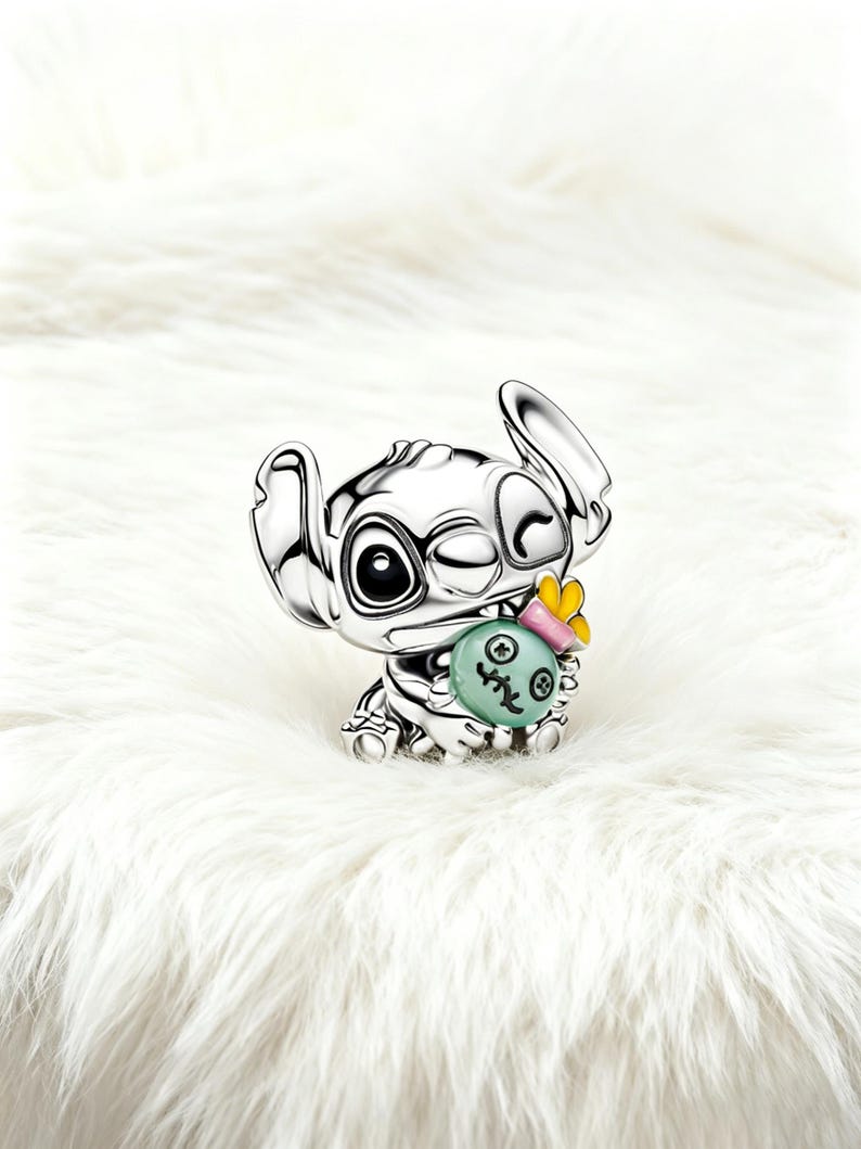 May include: A silver-colored figurine of the character Stitch from Lilo & Stitch. The figurine is winking and holding a light green object with a yellow bow. The figurine is on a white, fluffy surface.