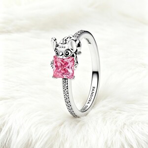 Puede incluir: Un anillo de plata con la figura esculpida de Stitch sosteniendo una piedra preciosa rosa de corte cuadrado. La banda est&aacute; decorada con peque&ntilde;as piedras transparentes en los laterales y grabada con PANDORA S925 y Disney en el interior.