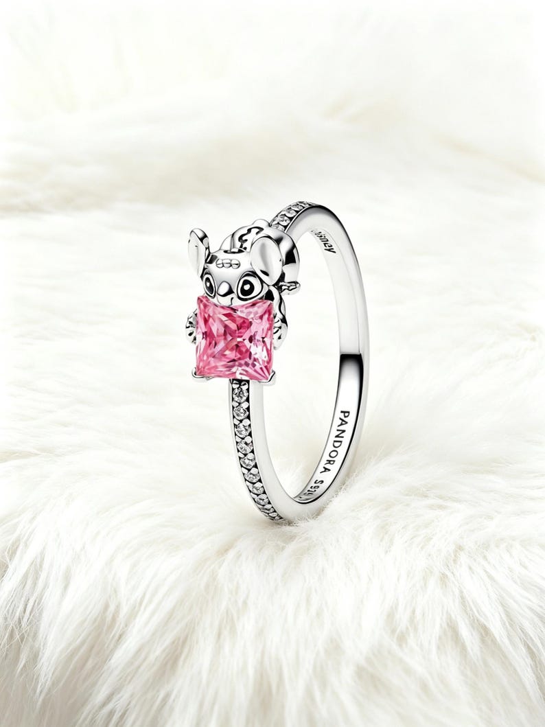 Puede incluir: Un anillo de plata con la figura esculpida de Stitch sosteniendo una piedra preciosa rosa de corte cuadrado. La banda est&aacute; decorada con peque&ntilde;as piedras transparentes en los laterales y grabada con PANDORA S925 y Disney en el interior.