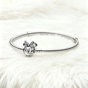 May include: A silver bracelet with a Minnie Mouse charm. The charm features a detailed Minnie Mouse face with a bow adorned with small, sparkling stones. The bracelet has a snake chain design and a secure clasp.