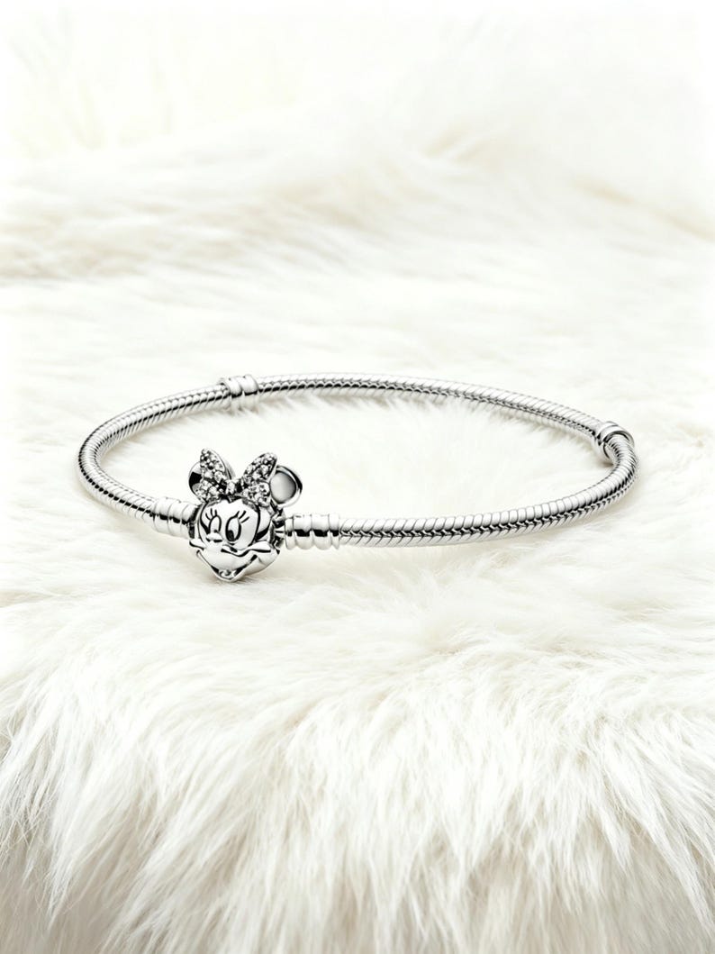 May include: A silver bracelet with a Minnie Mouse charm. The charm features a detailed Minnie Mouse face with a bow adorned with small, sparkling stones. The bracelet has a snake chain design and a secure clasp.