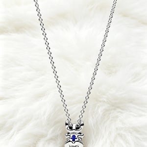 May include: A silver-colored necklace with a pendant shaped like a cartoon character. The pendant has a blue center and is suspended from a delicate chain. The necklace is displayed on a white, fluffy surface.