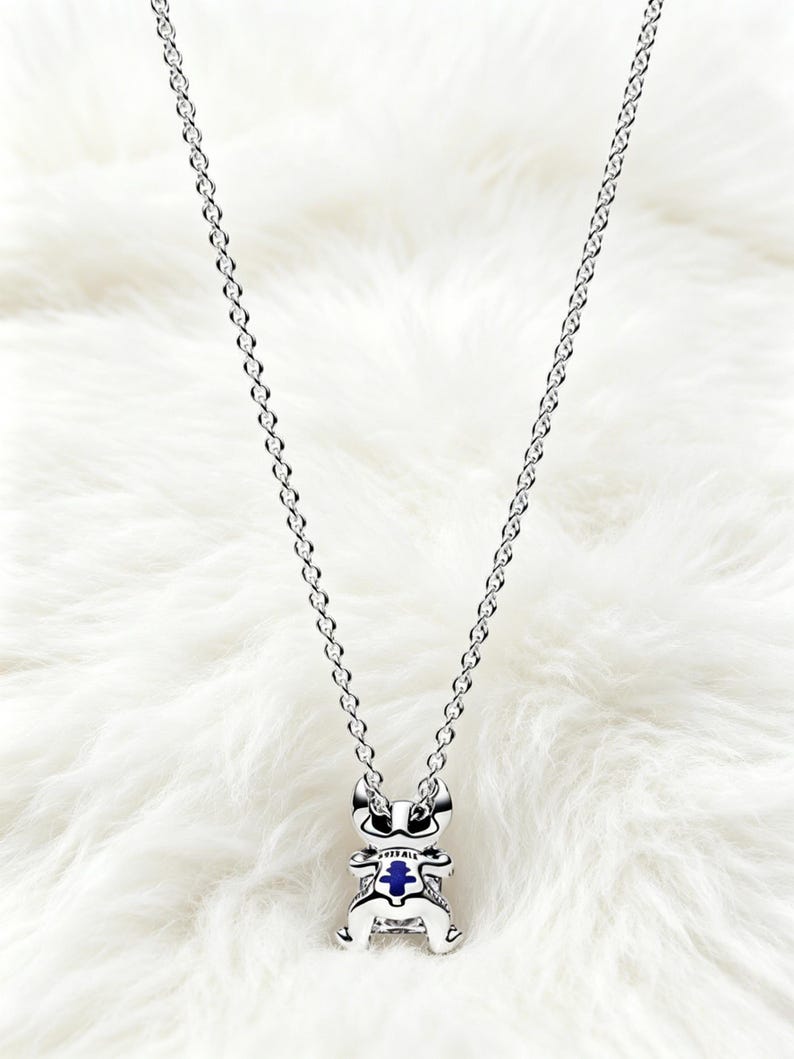 May include: A silver-colored necklace with a pendant shaped like a cartoon character. The pendant has a blue center and is suspended from a delicate chain. The necklace is displayed on a white, fluffy surface.