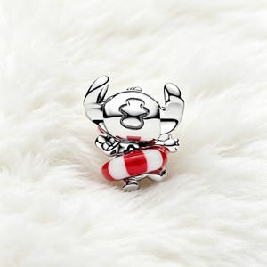 May include: A silver-colored charm depicting a cartoon character with large ears and a red and white striped floatation device. The charm is set against a white, fluffy background.