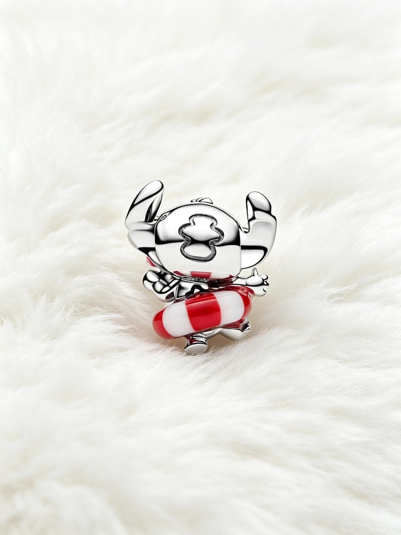 May include: A silver-colored charm depicting a cartoon character with large ears and a red and white striped floatation device. The charm is set against a white, fluffy background.