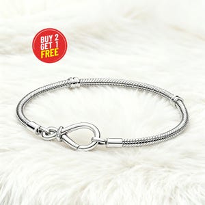 Könnte beinhalten: Ein silbernes Armband mit einem Unendlichkeitszeichen-Verschluss. Das Armband hat ein Schlangenketten-Design und wird auf einer weißen, flauschigen Oberfläche präsentiert. Eine rote und gelbe kreisförmige Grafik mit dem Text "BUY 2 GET 1 FREE" befindet sich oben links.