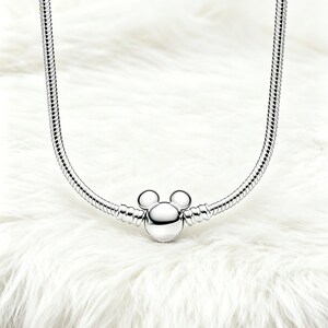 Peut inclure: Un collier argent&eacute; avec un charm Mickey Mouse. Le charm pr&eacute;sente une t&ecirc;te ronde et deux petites oreilles rondes. Le collier a un design de cha&icirc;ne serpent et est pr&eacute;sent&eacute; sur une surface blanche et duveteuse.