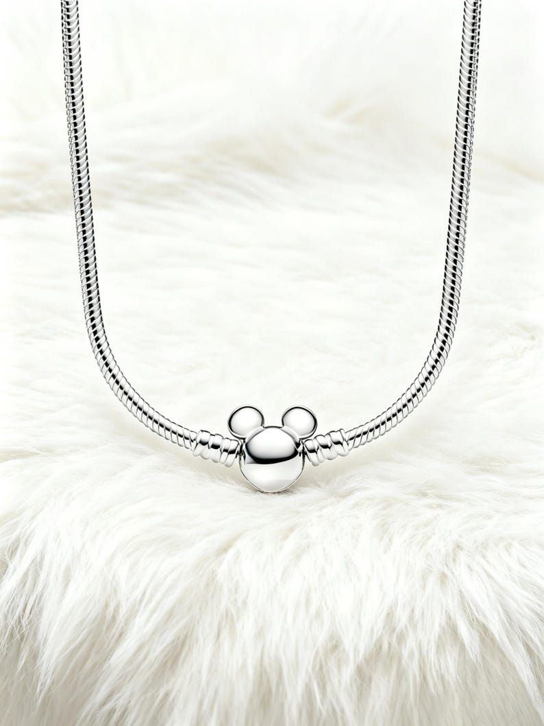 Peut inclure: Un collier argent&eacute; avec un charm Mickey Mouse. Le charm pr&eacute;sente une t&ecirc;te ronde et deux petites oreilles rondes. Le collier a un design de cha&icirc;ne serpent et est pr&eacute;sent&eacute; sur une surface blanche et duveteuse.