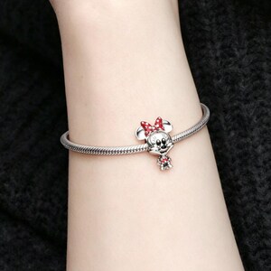 May include: A silver charm bracelet featuring a Minnie Mouse charm. The charm has a red bow and a small red dress. The bracelet is worn on a wrist, and the background is a dark, textured fabric.