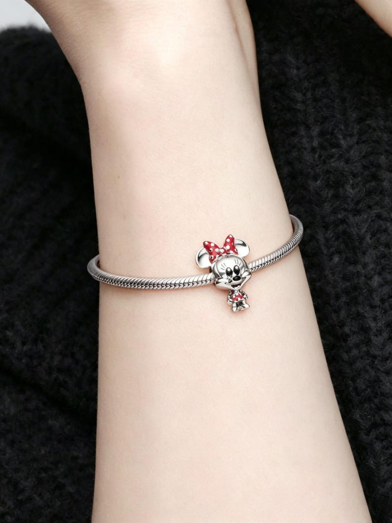 May include: A silver charm bracelet featuring a Minnie Mouse charm. The charm has a red bow and a small red dress. The bracelet is worn on a wrist, and the background is a dark, textured fabric.