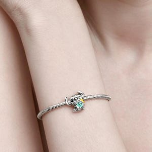 May include: A silver charm bracelet with a detailed charm of a cartoon character. The charm is a small, stylized figure with large eyes, ears, and a turquoise gem. The bracelet is worn on a wrist.