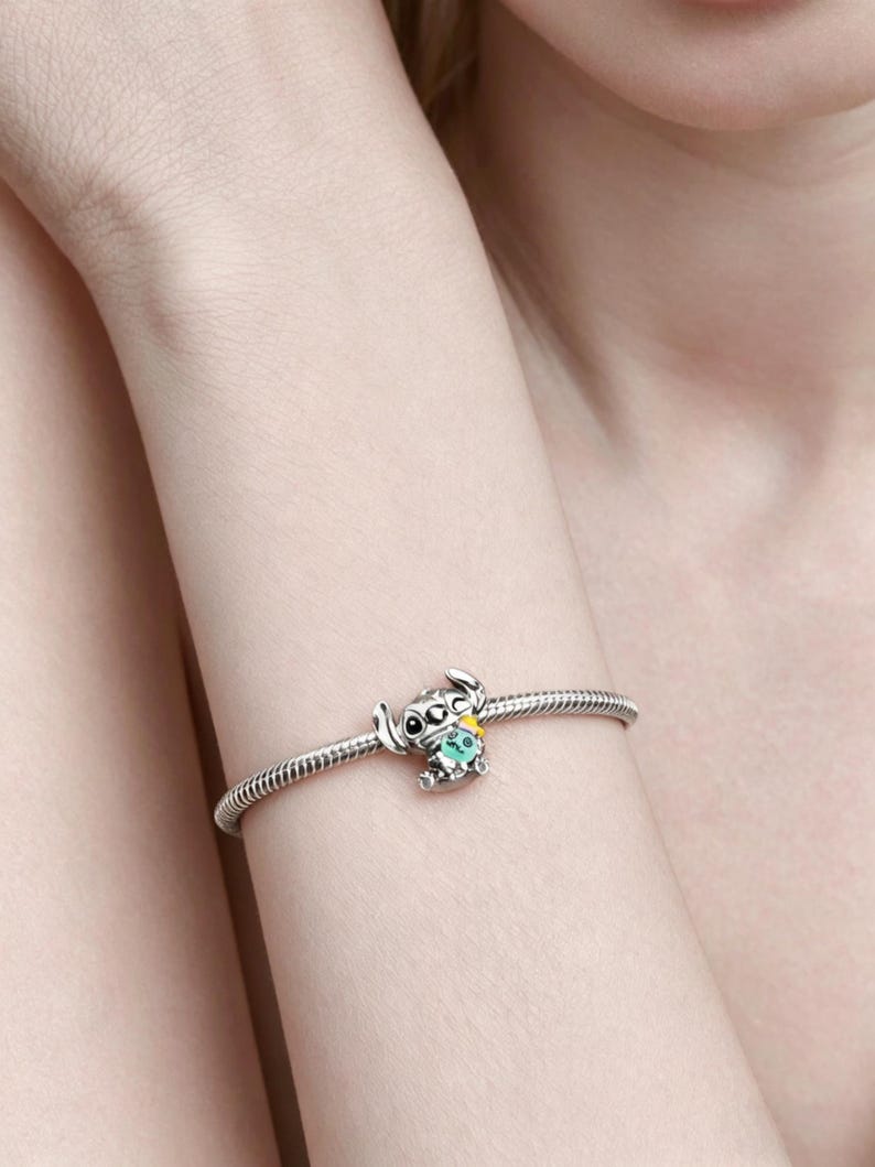 May include: A silver charm bracelet with a detailed charm of a cartoon character. The charm is a small, stylized figure with large eyes, ears, and a turquoise gem. The bracelet is worn on a wrist.