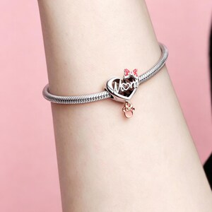 May include: Silver bracelet with a heart-shaped charm. The charm features the word "Mom" in script, a pink bow, and a small rose gold charm. The bracelet is on a person's wrist against a pink background.