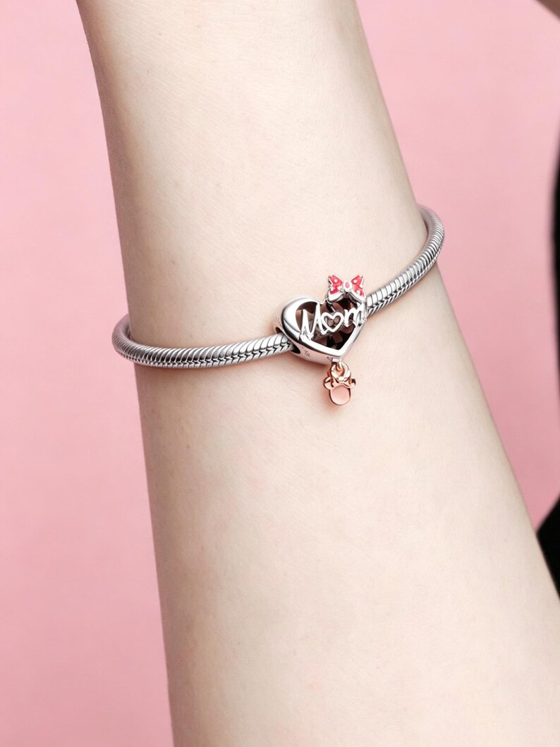 May include: Silver bracelet with a heart-shaped charm. The charm features the word "Mom" in script, a pink bow, and a small rose gold charm. The bracelet is on a person's wrist against a pink background.