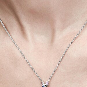 May include: A silver necklace with a pendant shaped like a cartoon character. The pendant features a clear, square-cut gemstone body, blue accents, and pink ears. The necklace is displayed against a skin-toned background.