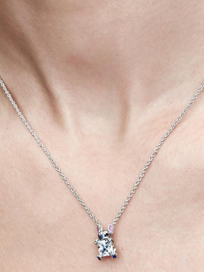 May include: A silver necklace with a pendant shaped like a cartoon character. The pendant features a clear, square-cut gemstone body, blue accents, and pink ears. The necklace is displayed against a skin-toned background.