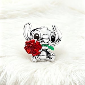 May include: A silver figurine of a cartoon character holding a red rose with a green stem. The character has large black eyes and large ears. The background is a white, fluffy surface.