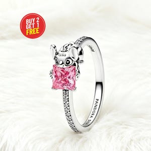 Puede incluir: Un anillo de plata con una figura que sostiene una piedra preciosa rosa de corte cuadrado. La banda est&aacute; incrustada con peque&ntilde;as piedras claras y grabada con el texto PANDORA S925 y Disney. Una insignia circular roja muestra BUY 2 GET 1 FREE.