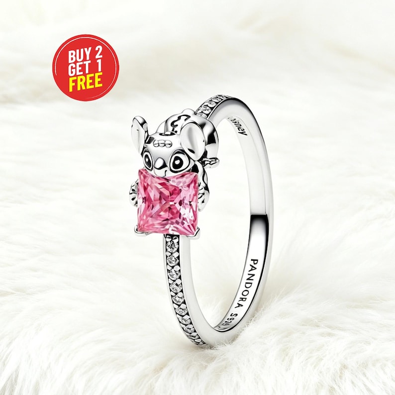 Puede incluir: Un anillo de plata con una figura que sostiene una piedra preciosa rosa de corte cuadrado. La banda est&aacute; incrustada con peque&ntilde;as piedras claras y grabada con el texto PANDORA S925 y Disney. Una insignia circular roja muestra BUY 2 GET 1 FREE.