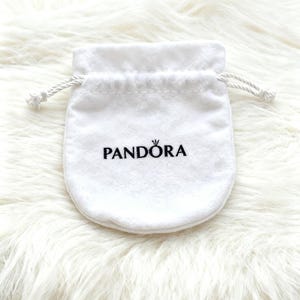 May include: A white jewellery pouch with a drawstring closure, featuring the black printed brand name "PANDORA". The pouch is made of a soft, velvety material and is set against a fluffy, white background.