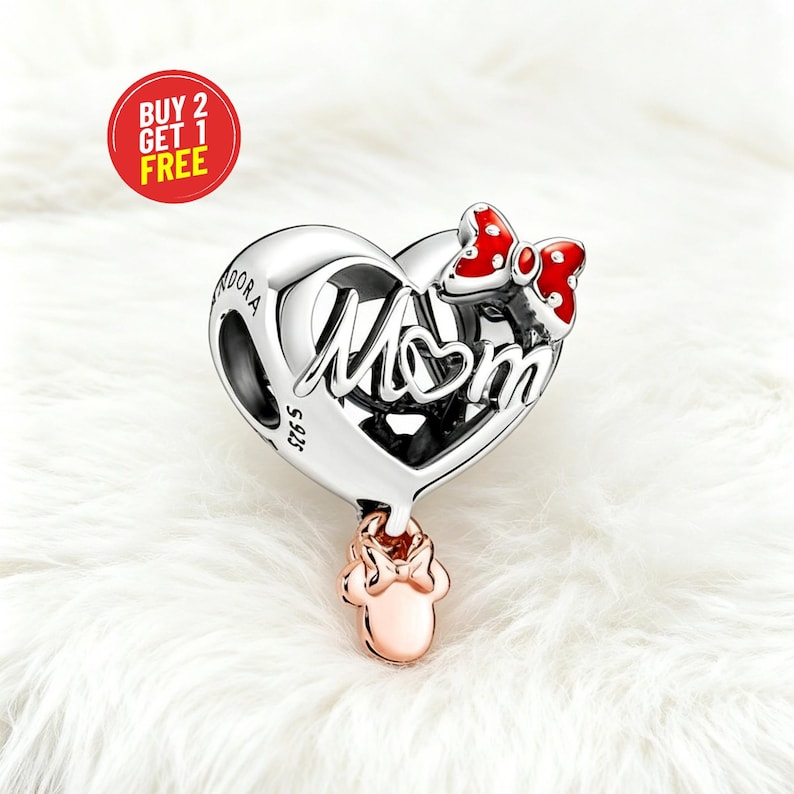 May include: A silver heart-shaped charm with the word "Mom" in cursive. A red bow with white polka dots is on the top right. A rose gold Minnie Mouse charm hangs below. The charm is on a white, fluffy surface.