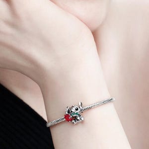 May include: Silver charm bracelet with a detailed Stitch charm holding a red heart. The charm has green accents and is attached to a silver chain bracelet. The bracelet is worn on a wrist.