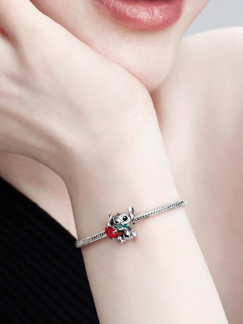 May include: Silver charm bracelet with a detailed Stitch charm holding a red heart. The charm has green accents and is attached to a silver chain bracelet. The bracelet is worn on a wrist.