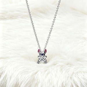 May include: A silver-toned necklace featuring a pendant shaped like a cartoon character. The pendant has a clear, faceted body, with pink, blue, and silver accents. The necklace hangs on a delicate chain against a white, fluffy background.