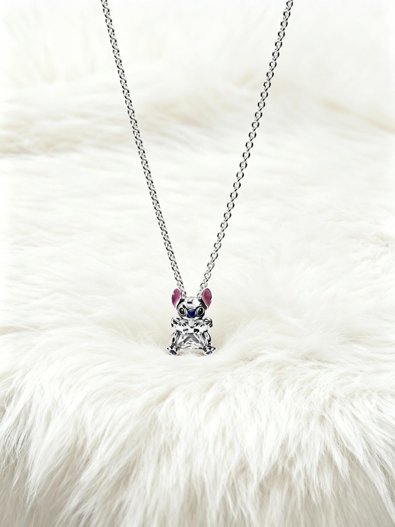 May include: A silver-toned necklace featuring a pendant shaped like a cartoon character. The pendant has a clear, faceted body, with pink, blue, and silver accents. The necklace hangs on a delicate chain against a white, fluffy background.