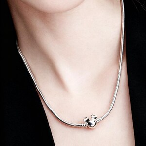 Peut inclure: Un collier argent&eacute; avec un charm Mickey Mouse. Le collier a un design de cha&icirc;ne serpent et un charm rond avec deux petits cercles sur le dessus, ressemblant aux oreilles de Mickey Mouse.