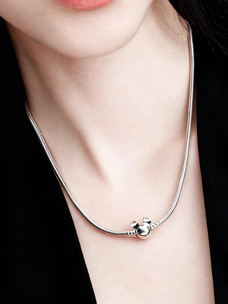 Peut inclure: Un collier argent&eacute; avec un charm Mickey Mouse. Le collier a un design de cha&icirc;ne serpent et un charm rond avec deux petits cercles sur le dessus, ressemblant aux oreilles de Mickey Mouse.