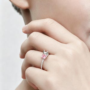 Puede incluir: Un anillo de tono plateado que presenta una peque&ntilde;a cabeza de personaje esculpida con orejas grandes sobre una piedra preciosa de color rosa claro y corte cuadrado. La banda es delgada y est&aacute; incrustada con una fila de piedras brillantes.