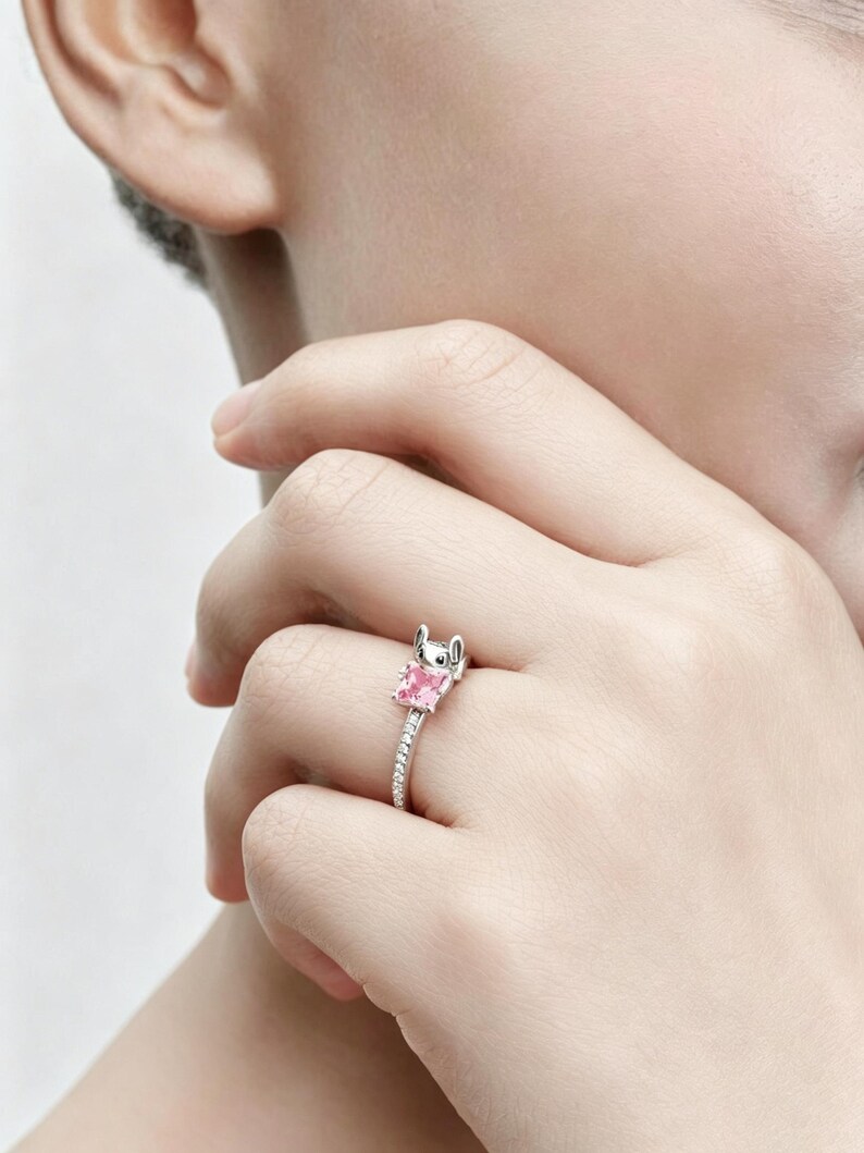 Puede incluir: Un anillo de tono plateado que presenta una peque&ntilde;a cabeza de personaje esculpida con orejas grandes sobre una piedra preciosa de color rosa claro y corte cuadrado. La banda es delgada y est&aacute; incrustada con una fila de piedras brillantes.