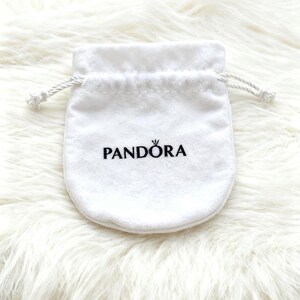 May include: A white, drawstring jewelry pouch with the brand name "PANDORA" printed in black. The pouch is made of a soft, velvety material and is set against a fluffy, white background.