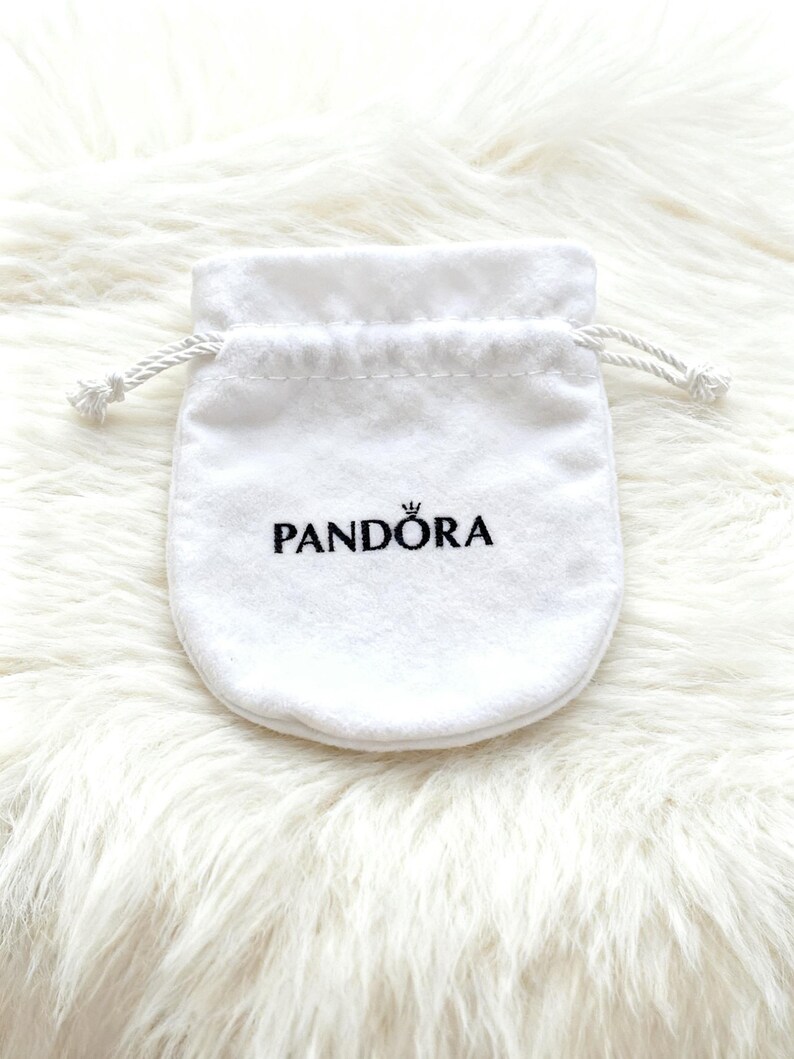 May include: A white, drawstring jewelry pouch with the brand name "PANDORA" printed in black. The pouch is made of a soft, velvety material and is set against a fluffy, white background.