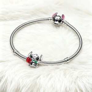 May include: A silver charm bracelet with two Stitch charms. One charm is a Stitch head with pink ears, and the other is Stitch holding a red rose. The bracelet is displayed on a white, fluffy surface.