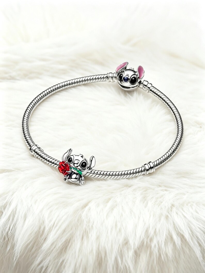 May include: A silver charm bracelet with two Stitch charms. One charm is a Stitch head with pink ears, and the other is Stitch holding a red rose. The bracelet is displayed on a white, fluffy surface.