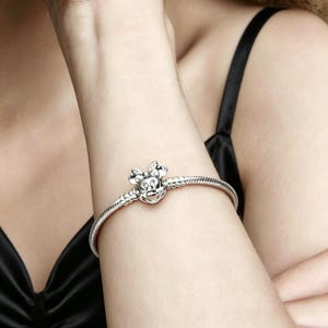 May include: A silver bracelet featuring a Minnie Mouse charm. The charm is detailed with a bow and face. The bracelet is worn on a person's arm, against a black top.