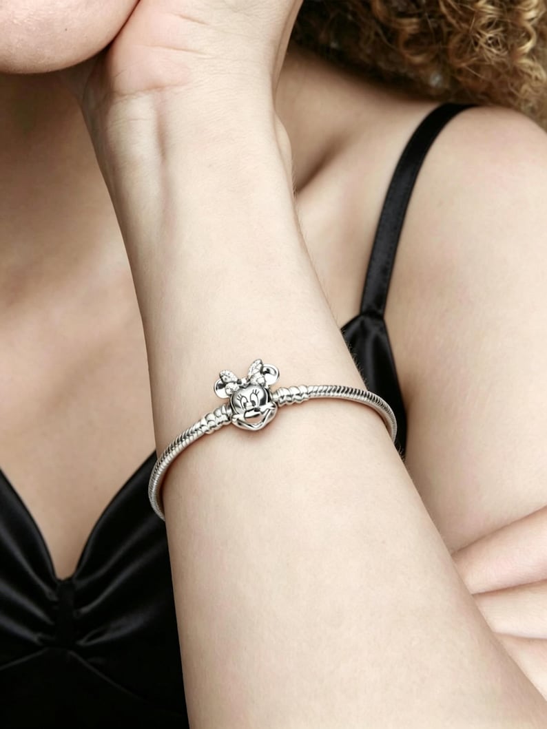 May include: A silver bracelet featuring a Minnie Mouse charm. The charm is detailed with a bow and face. The bracelet is worn on a person's arm, against a black top.