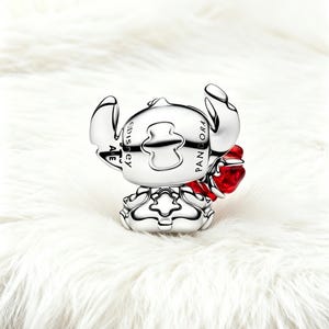 May include: A silver charm depicting the Disney character Stitch, with a red heart detail. The charm is engraved with "PANDORA" and "Disney". The charm is set against a white, fluffy background.