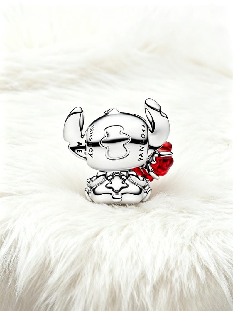 May include: A silver charm depicting the Disney character Stitch, with a red heart detail. The charm is engraved with "PANDORA" and "Disney". The charm is set against a white, fluffy background.