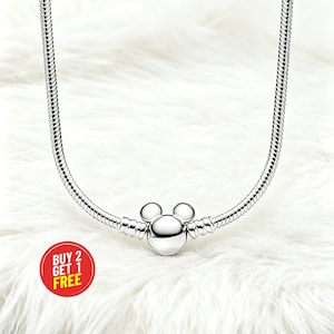 Peut inclure: Un collier argent&eacute; avec un charm Mickey Mouse. Le charm est au centre du collier et a une t&ecirc;te ronde avec deux petites oreilles rondes. Le collier est pos&eacute; sur une surface blanche et duveteuse. Un cercle rouge avec le texte "BUY 2 GET 1 FREE" est en bas &agrave; gauche.