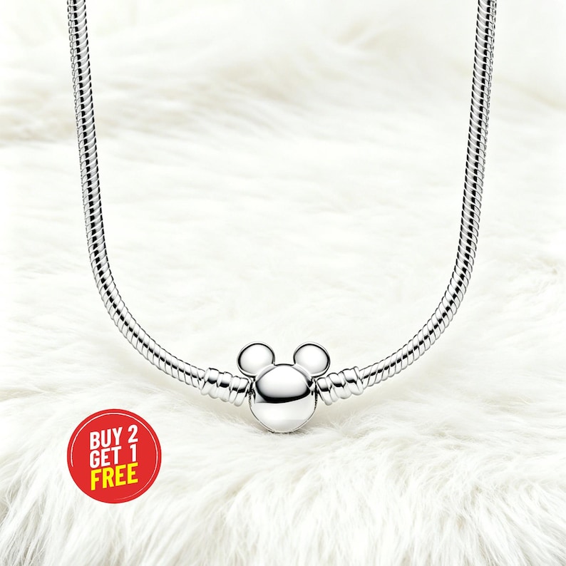 Peut inclure: Un collier argent&eacute; avec un charm Mickey Mouse. Le charm est au centre du collier et a une t&ecirc;te ronde avec deux petites oreilles rondes. Le collier est pos&eacute; sur une surface blanche et duveteuse. Un cercle rouge avec le texte "BUY 2 GET 1 FREE" est en bas &agrave; gauche.