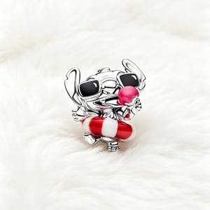 May include: A silver charm depicting a cartoon character wearing sunglasses and holding a pink ice cream cone. The character is also wearing a red and white striped life preserver. The charm is set against a white, fluffy background.