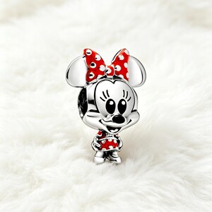 May include: A silver charm featuring Minnie Mouse with a red polka-dot bow and dress. The charm has a detailed design with black eyes, a black nose, and a smiling expression. The charm is a collectible item.