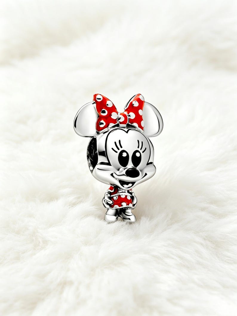May include: A silver charm featuring Minnie Mouse with a red polka-dot bow and dress. The charm has a detailed design with black eyes, a black nose, and a smiling expression. The charm is a collectible item.